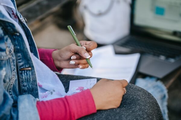 female student writing notes while sitting with laptop on bench
